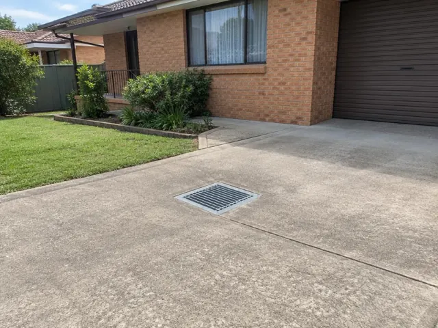 Concrete driveway with a square metal drain grate beside a brick house in Lalor Park