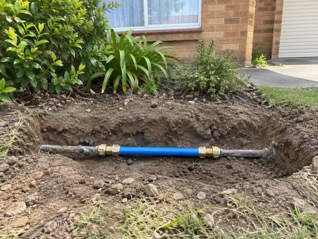 Open trench with a repaired blue water pipe section beside a brick house in Seven Hills
